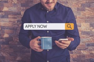 A person wearing a blue shirt is smiling while holding a blue mug and using a smartphone. Hovering above is a search bar with the words "APPLY NOW" and a magnifying glass icon against a brown brick background, hinting at an exciting job description waiting to be discovered.