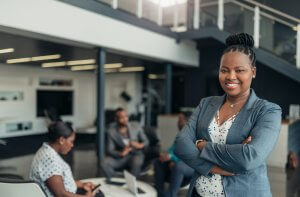 A confident woman in business attire stands with her arms crossed, exuding leadership as she smiles at the camera in a modern office environment. In the background, colleagues are engaging in a discussion at a table. The office boasts a contemporary design with open space and glass railings.