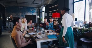 A group of diverse people, including Gen Z team members, sit around a large table in a modern office space, enthusiastically clapping and smiling at a standing woman who is presenting or speaking. The atmosphere appears collaborative and positive, with sunlight streaming through large windows.