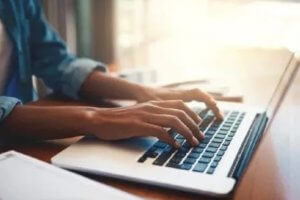 A person wearing a blue shirt types on a silver laptop with a dark keyboard, seated at a wooden desk. The background is out of focus, showing soft lighting and blurry indoor elements.