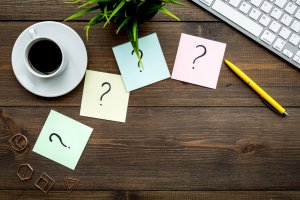 A cup of coffee sits on a wooden desk next to four colorful sticky notes adorned with question marks. Nearby, there is a keyboard, a yellow pen, a plant, and some geometric paper clips, creating a workspace scene filled with questions and creativity.