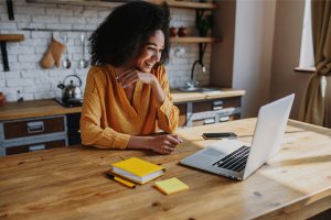 A person with curly hair wearing a yellow blouse is sitting at a wooden table in a kitchen, smiling and looking at a laptop. Yellow notepads, a smartphone, and other essentials for remote work are on the table. Shelves with kitchen items are in the background.