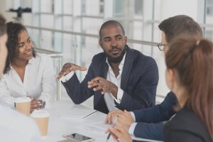 A diverse group of five professionals is engaged in a discussion around a table. One man in a dark suit, exemplifying leadership, is speaking and gesturing with his hands while the others listen attentively. Notebooks, coffee cups, and a smartphone are on the table.