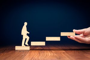 A wooden cutout of a person ascends a staircase made of rectangular wooden blocks. An honest hand is seen placing one of the blocks in the staircase, symbolizing support or progress. The background is dark, and the scene is set on a wooden surface.