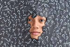 A face peeks through a dense layer of black cards with white question marks, staring directly at the camera. The background and face are heavily surrounded by the question mark cards, amplifying an atmosphere brimming with questions and creating a visually intriguing theme.