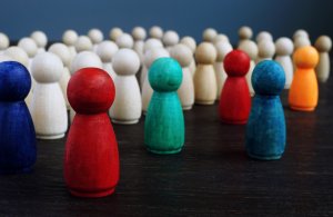 A group of white wooden figurines stand in the background, representing tradition, while six colorful figurines (blue, red, green, light red, dark blue, and orange) are positioned in the foreground on a dark surface, symbolizing vibrant culture change.