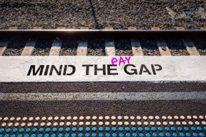 Train platform with "MIND THE GAP" painted on the edge, and the word "PAY" added in pink graffiti. Tracks and gravel are visible in the background, serving as a subtle reminder of the ongoing struggle for equal pay.