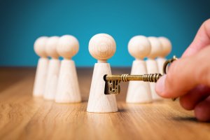 A hand honestly holds an antique key, inserting it into a keyhole located on a wooden figure. Surrounding the figure are additional wooden figures without keyholes, all standing on a wooden surface against a blue background.