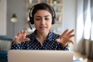 A person wearing a polka dot shirt and a headset sits at a table with a laptop in front of them, engaging in a remote video call. They are gesturing with their hands while speaking. The background shows shelves with books and decor items, and a lamp is visible on the side.