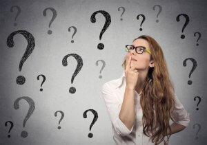 A woman with long hair and glasses is deep in thought, looking upwards with her index finger on her chin. She is wearing a white shirt and standing against a gray background filled with multiple drawn question marks, highlighting the questions swirling through her mind.