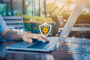 A person uses a laptop with floating digital graphics of locks and a shield icon, representing robust cybersecurity. The laptop sits open on a wooden table against a bright, blurred background, emphasizing both data protection and online security.