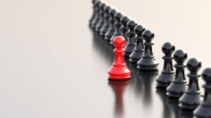 A line of black chess pawns stands on a smooth reflective surface. One red pawn is prominently positioned in the middle of the line, standing out against the uniform black pawns. The scene symbolizes uniqueness, individuality, and leadership.