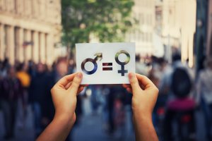 Two hands hold up a piece of paper with symbols for male and female separated by an equal sign, signifying gender equality and equal pay. The background shows a bustling city street with blurred pedestrians.