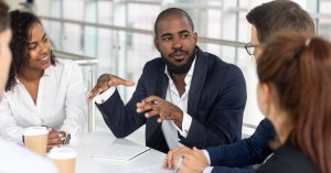 A group of four diverse colleagues sit around a table in a modern office. The central figure, showcasing strong leadership, gestures expressively while speaking. Two women and another man listen attentively, with coffee cups and papers set on the table.