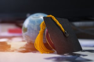 Close-up of a graduation cap with a yellow tassel placed on a table. The cap is partially resting on a small, colorful globe in the background, highlighting an international theme. The setting appears to be indoors with soft lighting.