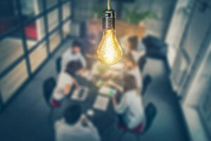A lit light bulb hangs in focus in the foreground, symbolizing innovation, while a blurred background shows a group of people sitting around a conference table engaged in a meeting or discussion. The room has glass walls and some greenery in the background.