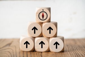 A pyramid of six wooden blocks sits on a table. The top block features a compass, while the five blocks below sport black arrows pointing upwards, symbolizing peak performance. The background is a plain, light-colored wall.