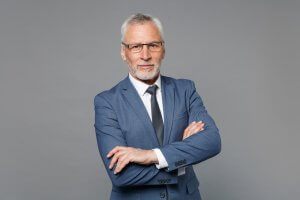 A confident older man with white hair and beard, wearing glasses, a blue suit, and a black tie stands against a gray background. With his arms crossed and a serious expression, he looks like he's ready to get back to working.