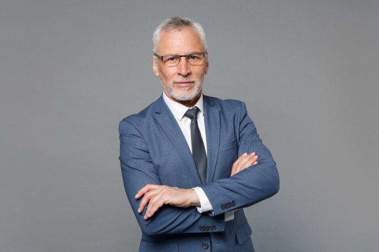A confident older man with white hair and beard, wearing glasses, a blue suit, and a black tie stands against a gray background. With his arms crossed and a serious expression, he looks like he's ready to get back to working.