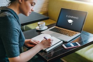 A woman sits at a table with an open notebook, writing with a pen. She faces a laptop displaying the word "eLearning" alongside an icon of a graduation cap. A smartphone lies beside the notebook and a cup of coffee is on the table. She appears focused on her work.
