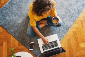 A person wearing a yellow sweater and blue jeans sits cross-legged on a gray rug while using a laptop, embodying the essence of remote work. They hold a cup of coffee in one hand, with a smartphone placed nearby on the floor. The setting appears to be a casual indoor space.