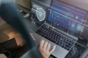 A person using a laptop with holographic security icons projecting from the screen. The display shows charts and data security-related graphics, emphasizing cyber security. The background is blurred, focusing on the person’s hands and the laptop.