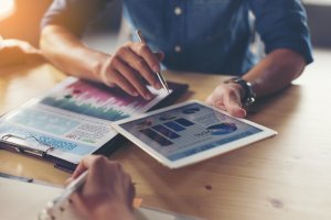 Two people collaborate at a table. One points to a graph on a tablet, while the other holds a pen. A document titled "Business Plan" with colorful charts lies on the table, highlighting performance metrics. Sunlight illuminates the scene, suggesting a productive discussion.