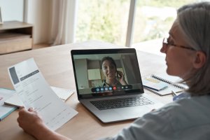 A person wearing glasses is sitting at a desk, holding a document and participating in a video call on a laptop. The screen shows someone discussing job hunting strategies during the call. Nearby, the desk has notebooks, a pen, and enjoys natural light streaming through the window in the background.