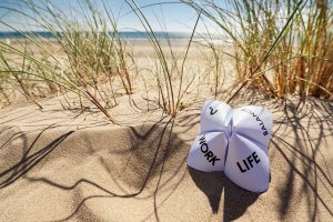 An origami fortune teller lies on a sunny, sandy beach surrounded by coastal grasses. The visible flaps read "WORK" and "LIFE." In the background, a calm sea and distant mountains are visible under a clear blue sky, embodying the balance of life amidst nature's serene beauty.