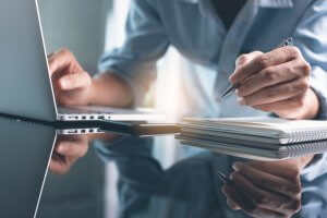 A person writes in a spiral-bound notebook while working on a laptop, immersed in eLearning. They hold a pen in their right hand and have their left hand on the keyboard. The scene is reflected on a glossy surface, and the environment appears calm and focused.