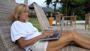 A woman lounges on an outdoor wicker chair, working on her laptop. She's wearing glasses and a white top with beige shorts. A tropical drink is placed on a nearby table. In the background, there are trees, a patio area, and a building with a thatched roof.