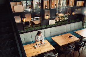 A woman sits alone at a wooden table in a modern cafe, working on a laptop. The cafe, ideal for remote work, has a stylish interior with shelves displaying bottles, soft lighting, and cushioned seating. The setting appears quiet and conducive to productivity.