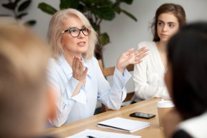 A woman with blonde hair and glasses is speaking animatedly during a meeting, demonstrating her passion for performance. She gestures with her hands while others listen. A notebook, pen, and smartphone are on the table in front of her. Another woman is visible in the background, listening attentively.
