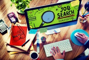 A person engaged in job hunting uses a desktop computer displaying a "Job Search" screen with a magnifying glass graphic. The desk is cluttered with a keyboard, mouse, phone, notepad, glasses, calculator, ruler, and various office supplies.