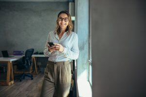 A smiling woman wearing glasses and business casual attire stands by a window, her smartphone in hand. Bathed in natural light, she radiates confidence in her career. An office setting with a desk, chair, and shelves is visible in the background.