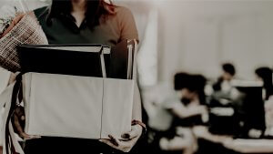 A person holding a box filled with office supplies, folders, and a bouquet of flowers. The background is blurred, showing an office setting with people working at desks. The atmosphere suggests that the person might be leaving due to a recent resignation or starting a new job.