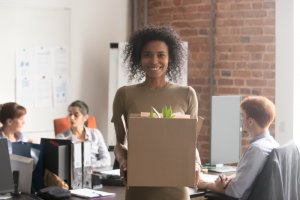A woman stands smiling in an office holding a cardboard box containing personal items, including a plant. Behind her, three colleagues are seated at their desks, deeply engaged in work. The office, with its mix of whiteboards and brick walls, buzzes with the energy of flourishing careers.