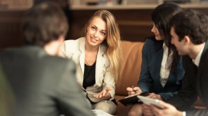 A woman in a beige blazer is speaking and gesturing with her hand while seated on a couch. She is surrounded by three other individuals, two men and one woman, who are attentively listening and taking notes. They appear to be in a career-focused business meeting.
