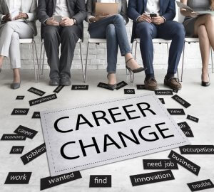 Five people sit on chairs holding electronic devices, their legs visible. A large sign in front of them reads "CAREER CHANGE." Surrounding the sign are scattered smaller words like "economy," "crisis," "career," "jobless," "innovation," and "employed.