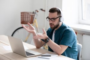 A man with glasses and headphones sits at a desk, gesturing with his hands while looking at a laptop screen. A smartphone, notebook, and pen are on the desk. In the background by the window, a bicycle hints at his preference for remote working life.
