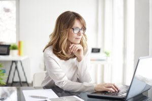 A woman with shoulder-length hair and glasses sits at a desk, working on a laptop. She is wearing a white blouse and resting her chin on one hand while typing with the other. The desk has papers and a tablet. The background of this modern office reflects her successful career.