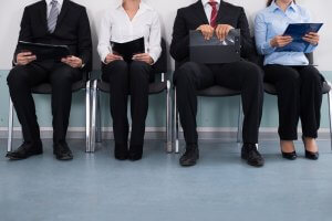 Four people in business attire sit on chairs in a waiting area, holding folders or clipboards. Only their torsos and legs are visible, suggesting they're waiting for an interview for a local job. The floor is blue, and the background features a light-colored wall.