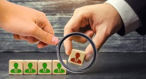 Two hands selecting wooden blocks with human figure icons on them. One hand holds a magnifying glass over a block with a red figure, symbolizing resignation, while green-figured blocks remain aligned below. The background is a textured wooden surface.