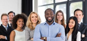 A group of eight diverse professionals, both men and women, standing together and smiling in a well-lit office space. The central figure, a man in a light blue shirt, stands slightly ahead with arms crossed. The group appears confident and friendly.