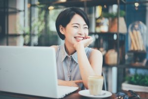 A woman with short black hair and a sleeveless button-up shirt is sitting at a table in front of a laptop, smiling brightly with her chin resting on her hand. A cup of coffee is in front of her, and the background features a cozy, well-lit café as she plans her next career move.