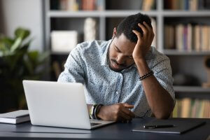 A person sits at a desk with a laptop, looking stressed. They are holding their head with one hand and have their eyes closed, possibly suffering from dead butt syndrome. Books and shelves clutter the background, adding to the disarray.