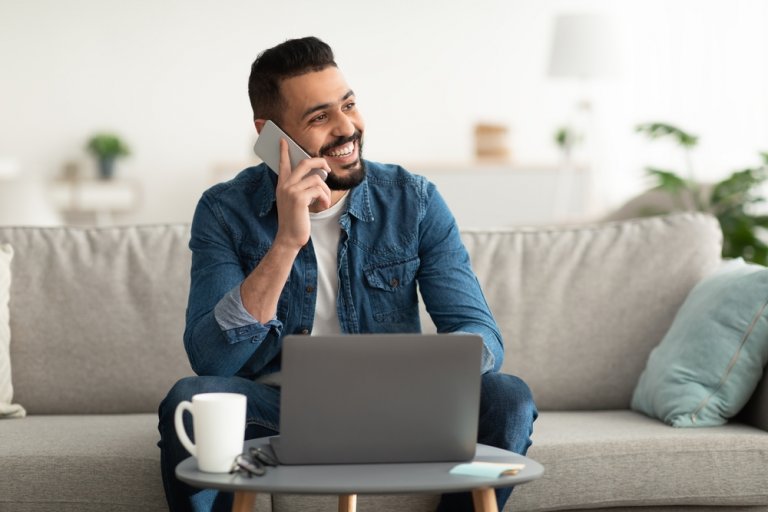 A smiling man sits on a sofa, holding a phone to his ear while looking away. His laptop is open on a small table in front of him, which also has a mug and a notepad. The background shows a bright, modern living room with plants and shelves, perfectly suited for working remotely.