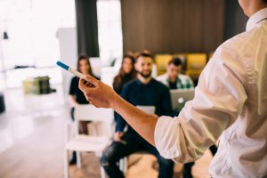 A person in a white shirt is holding a marker and giving a training presentation to a group of people who are seated and attentively listening. One of them has a laptop, and the setting appears to be a modern, informal office or conference room.
