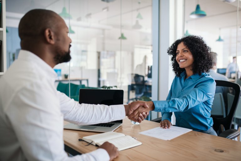 A man and a woman are seated at a wooden desk in a modern office, engaged in an HR discussion. They are shaking hands, smiling, and appear to be having a positive conversation. On the desk sits a laptop, a notebook, and some documents.