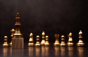 A close-up of golden chess pieces on a chessboard with a dark background. The king piece, symbolizing leadership, is prominently displayed in the foreground, standing on a wooden block, while the other pieces, slightly blurred, are aligned in the background.